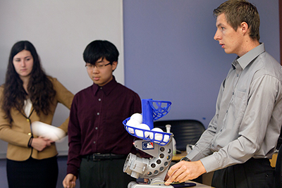 Margaret Grasse and Luolei Zhao look on as Brandon Tanner uses the team's Whistle Ball Launcher to shoot a ball across the room