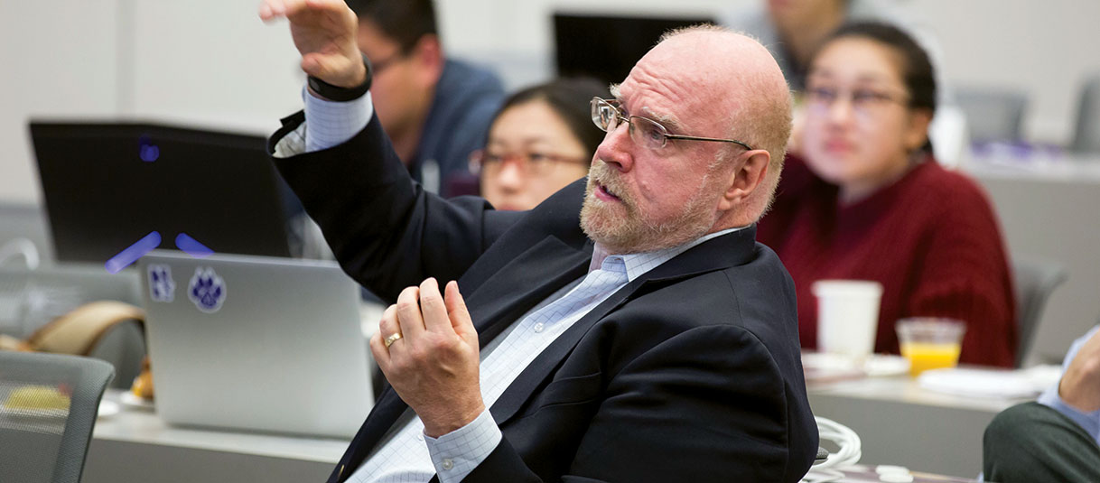 Faculty member sitting in a chair and surrounded by students in a classroom